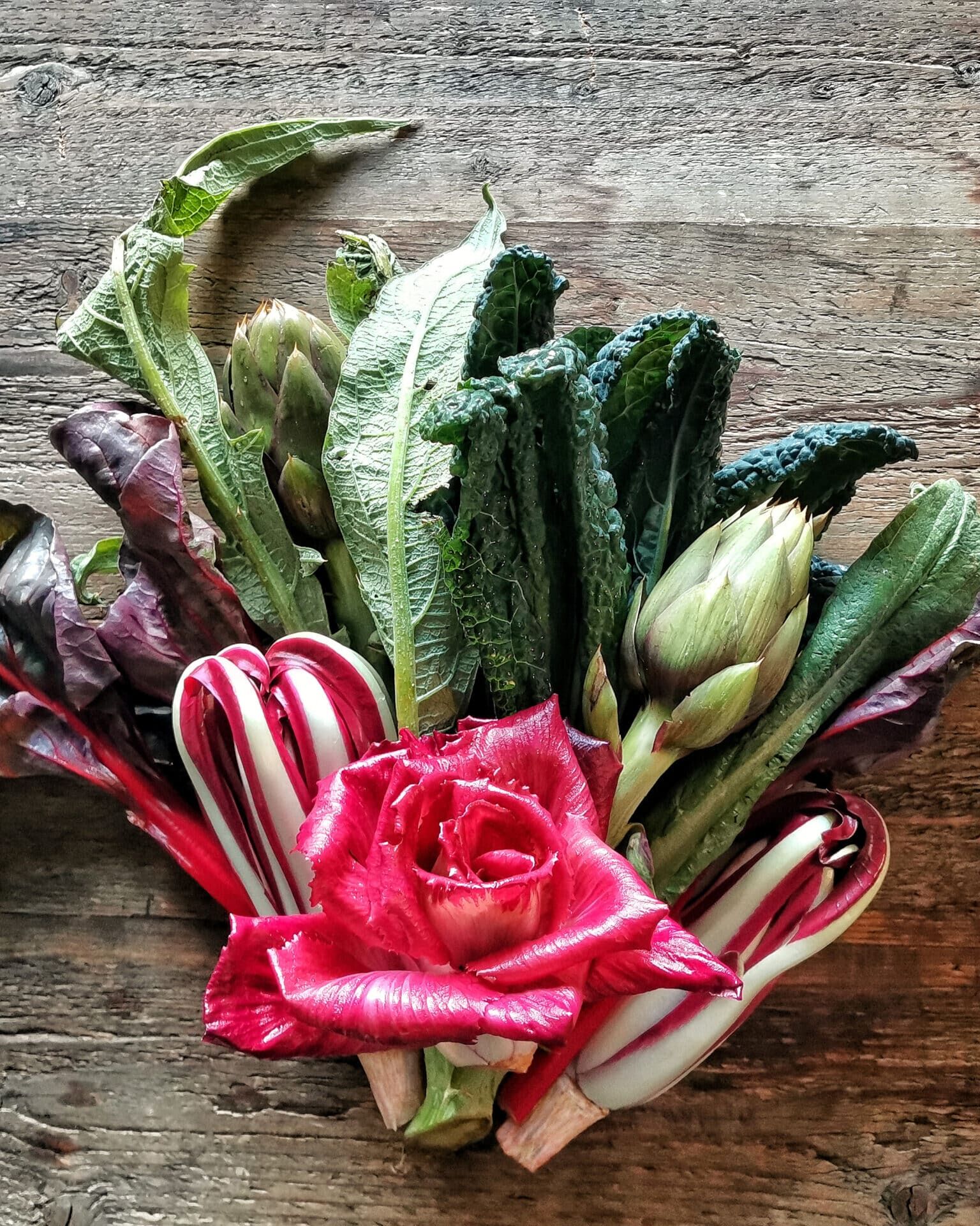 Fumagalli Danilo Ingrosso Ortofrutta Un mazzo di verdure a foglia verde assortite, due carciofi e gambi di bietola rossa disposti attorno a una singola rosa rossa su una superficie rustica in legno: l’ispirazione perfetta per chi opera nel settore della ristorazione o è alla ricerca di prodotti di prima scelta presso un grossista ortofrutticolo. Ristorazione Professionale - GDO - retail - privati