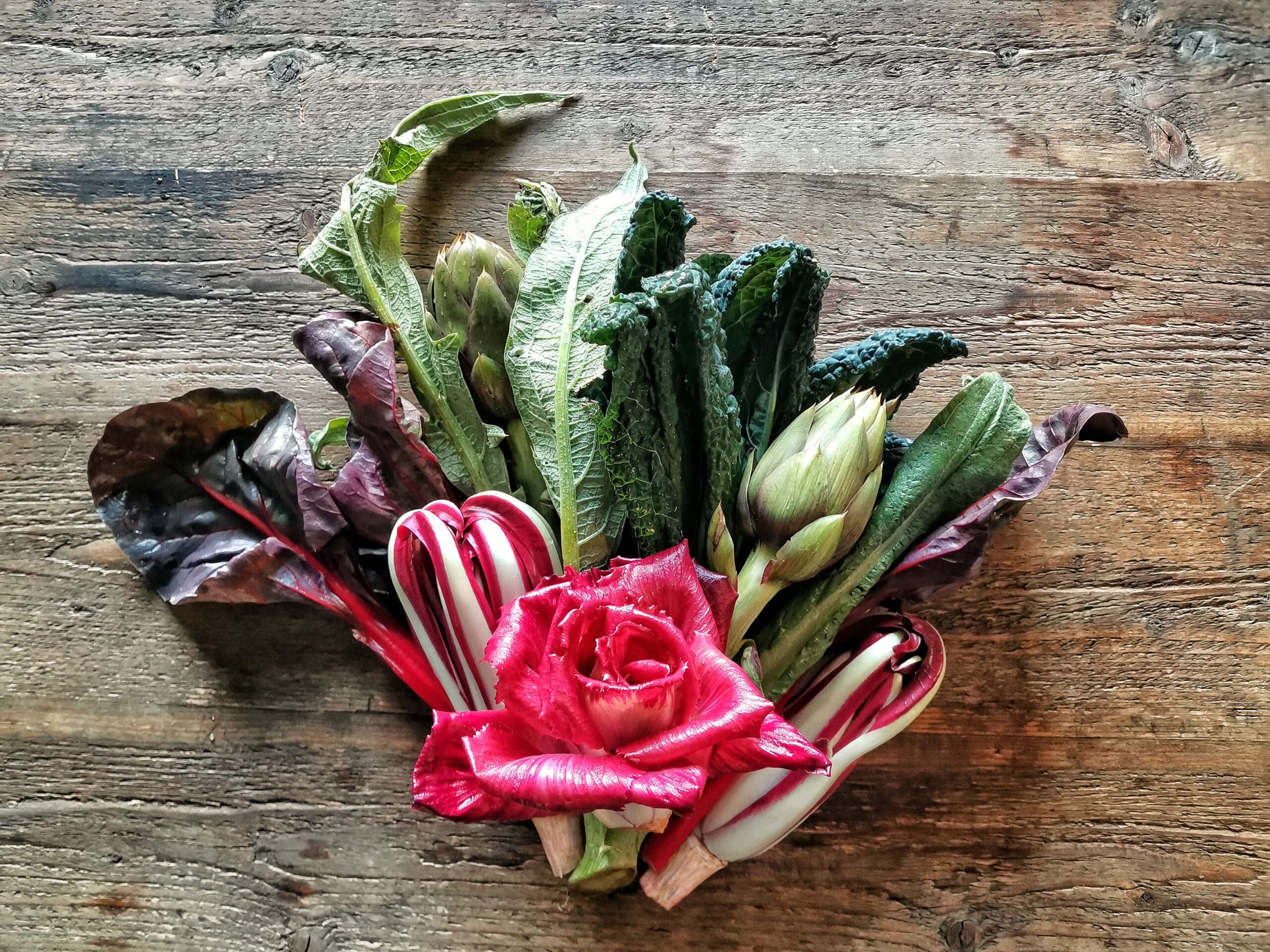 Fumagalli Danilo Ingrosso Ortofrutta Un mazzo di verdure a foglia verde assortite, due carciofi e gambi di bietola rossa disposti attorno a una singola rosa rossa su una superficie rustica in legno: l&rsquo;ispirazione perfetta per chi opera nel settore della ristorazione o &egrave; alla ricerca di prodotti di prima scelta presso un grossista ortofrutticolo. Ristorazione Professionale - GDO - retail - privati