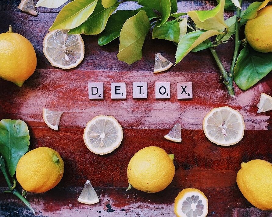 Fumagalli Danilo Ingrosso Ortofrutta Limoni interi e a fette, con le foglie, sono disposti attorno alle tessere dello Scrabble che compongono la parola “DETOX” su una superficie di legno, mettendo in risalto la freschezza che ci si aspetta da un grossista ortofrutticolo come Fumagalli Danilo Besana in Brianza. Ristorazione Professionale - GDO - retail - privati