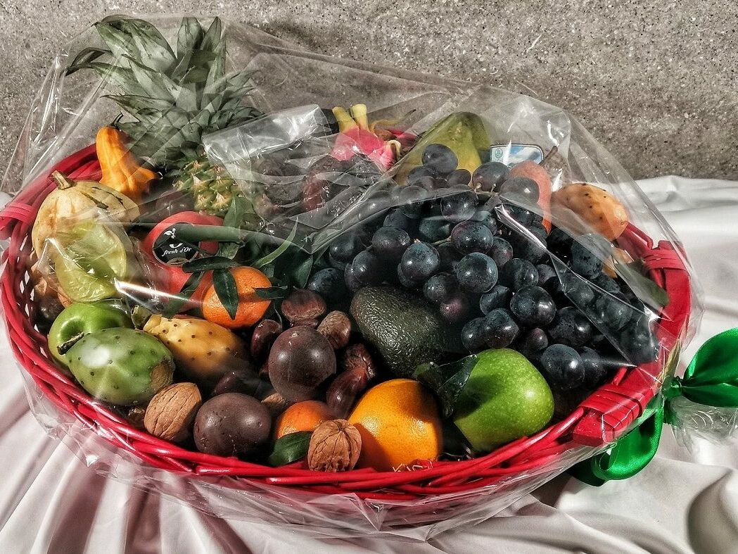 Fumagalli Danilo Ingrosso Ortofrutta Su una tovaglia bianca è posato un cesto rosso pieno di frutta e frutta secca assortite, provenienti da un grossista ortofrutta, avvolto in pellicola trasparente e legato con un nastro verde. Ristorazione Professionale - GDO - retail - privati