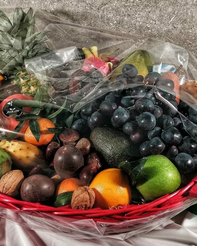 Fumagalli Danilo Ingrosso Ortofrutta Su una tovaglia bianca è posato un cesto rosso pieno di frutta e frutta secca assortite, provenienti da un grossista ortofrutta, avvolto in pellicola trasparente e legato con un nastro verde. Ristorazione Professionale - GDO - retail - privati