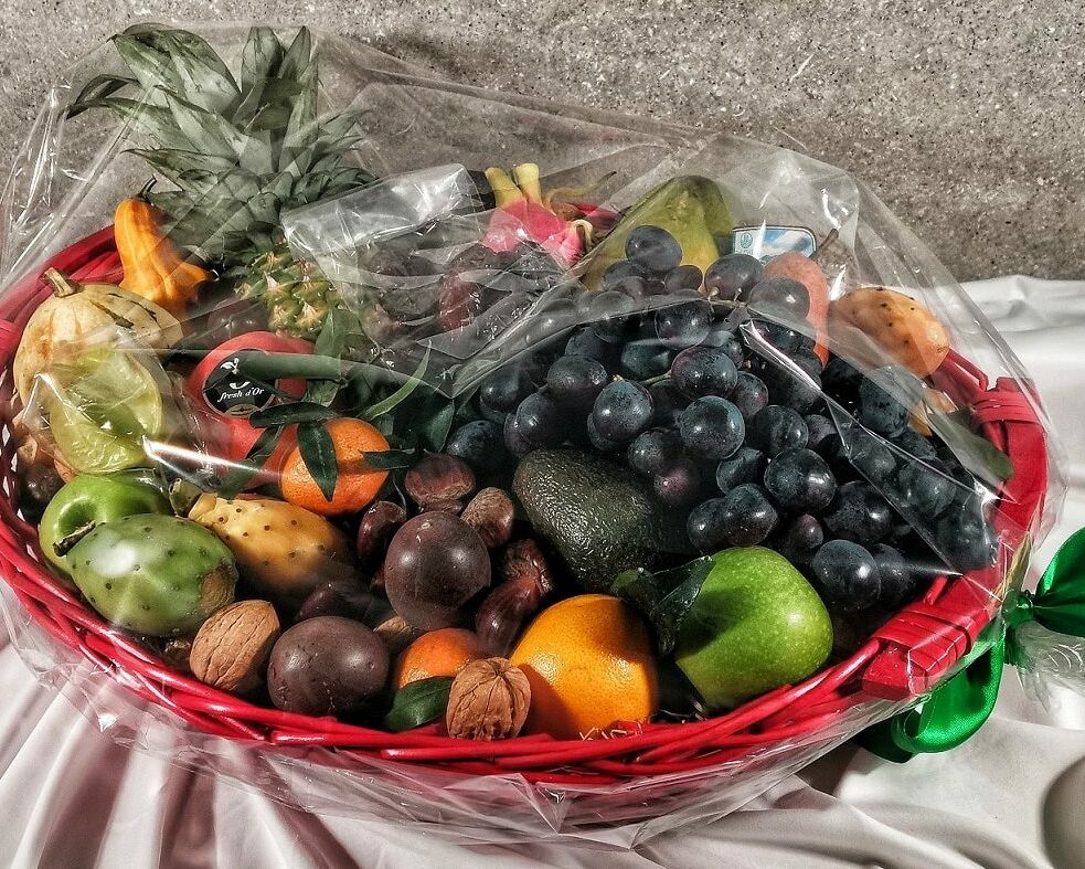 Fumagalli Danilo Ingrosso Ortofrutta Su una tovaglia bianca è posato un cesto rosso pieno di frutta e frutta secca assortite, provenienti da un grossista ortofrutta, avvolto in pellicola trasparente e legato con un nastro verde. Ristorazione Professionale - GDO - retail - privati
