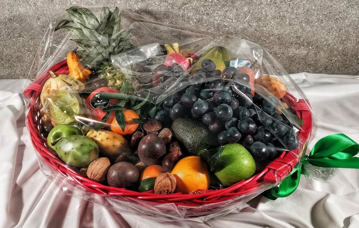 Fumagalli Danilo Ingrosso Ortofrutta Su una tovaglia bianca è posato un cesto rosso pieno di frutta e frutta secca assortite, provenienti da un grossista ortofrutta, avvolto in pellicola trasparente e legato con un nastro verde. Ristorazione Professionale - GDO - retail - privati