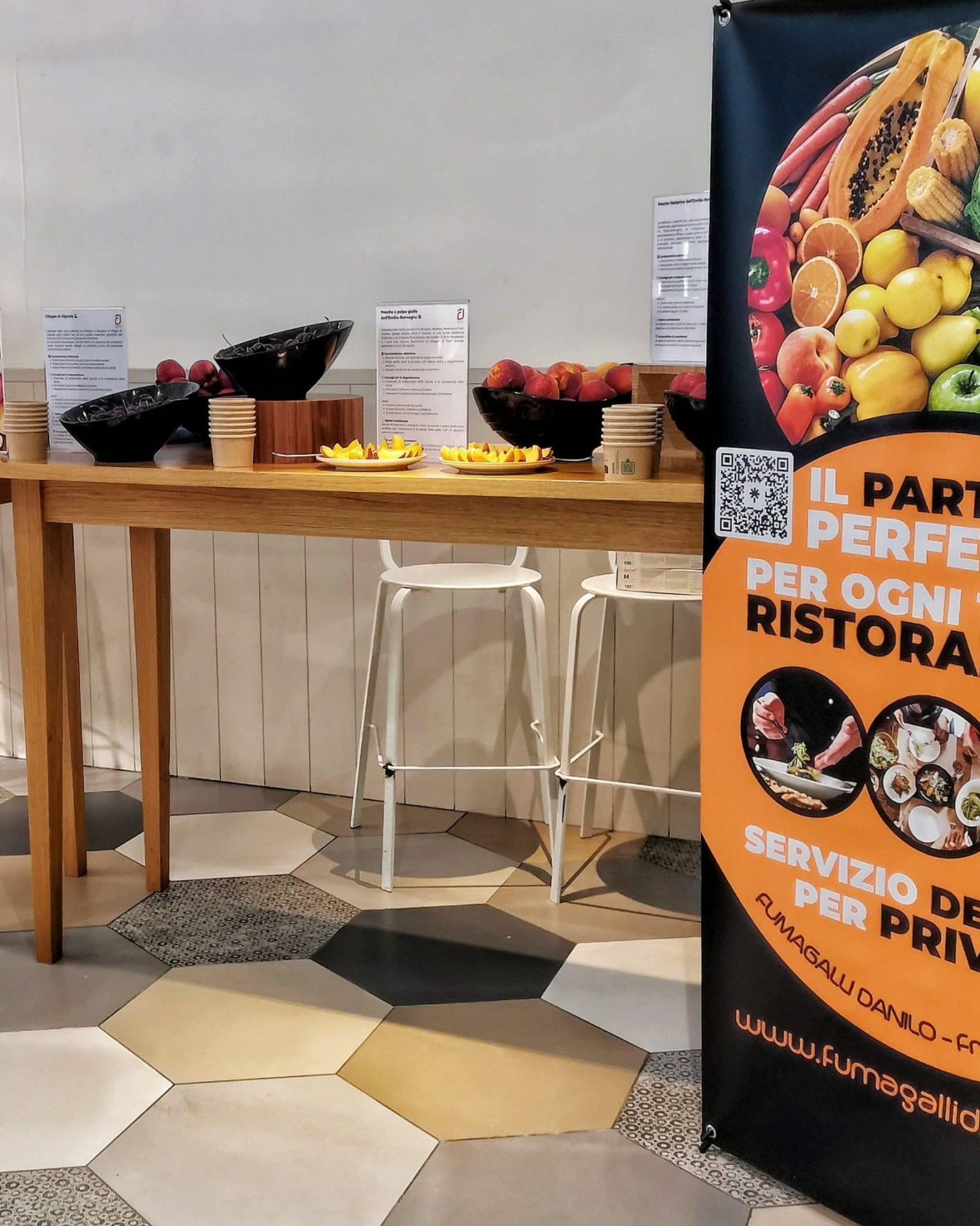 Fumagalli Danilo Ingrosso Ortofrutta Una donna è seduta a un tavolo alto su cui sono disposte delle ciotole di frutta; uno striscione colorato pubblicizza un servizio di vendita all’ingrosso di frutta e verdura per ristoranti e clienti privati. Ristorazione Professionale - GDO - retail - privati