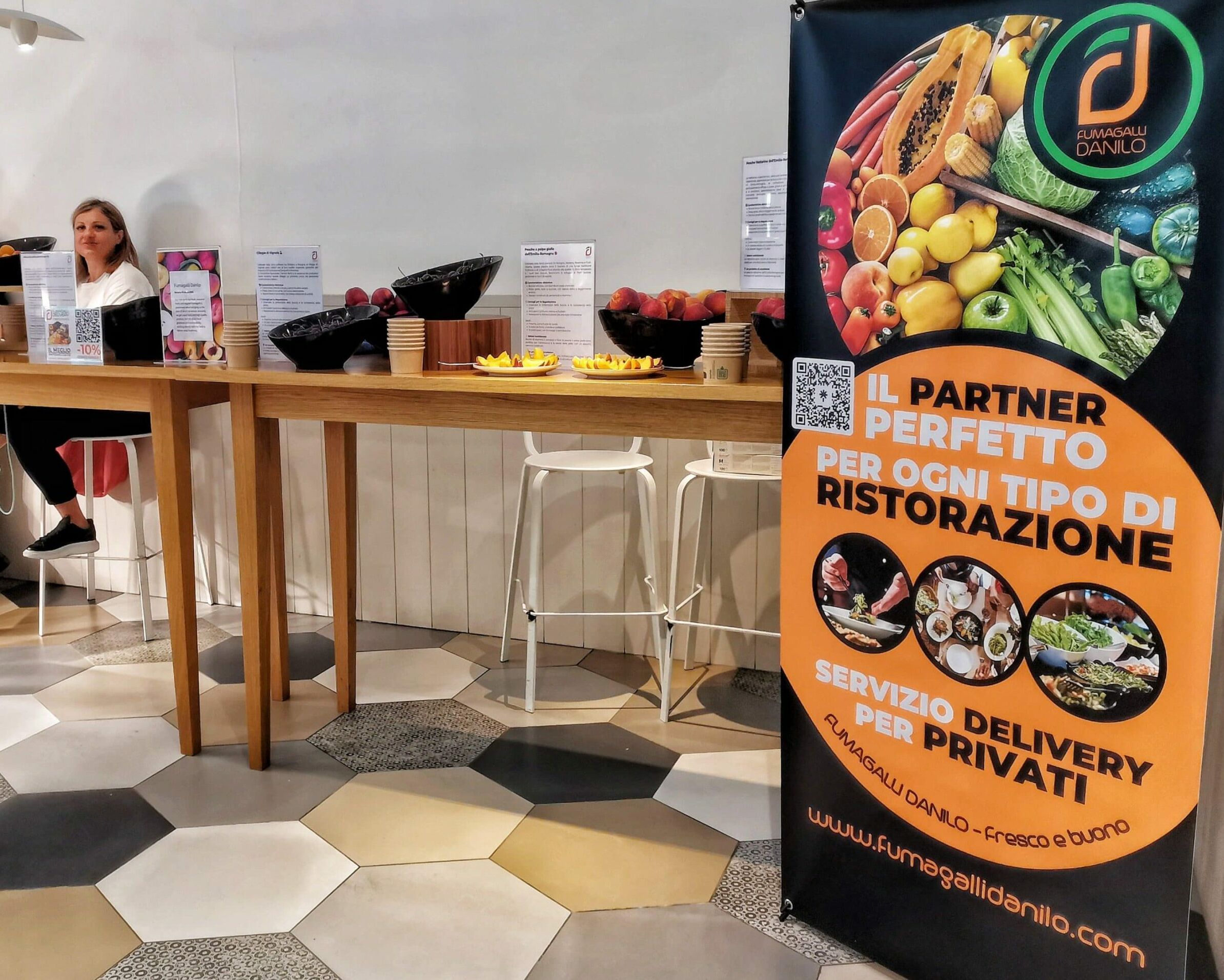 Fumagalli Danilo Ingrosso Ortofrutta Una donna è seduta a un tavolo alto su cui sono disposte delle ciotole di frutta; uno striscione colorato pubblicizza un servizio di vendita all’ingrosso di frutta e verdura per ristoranti e clienti privati. Ristorazione Professionale - GDO - retail - privati