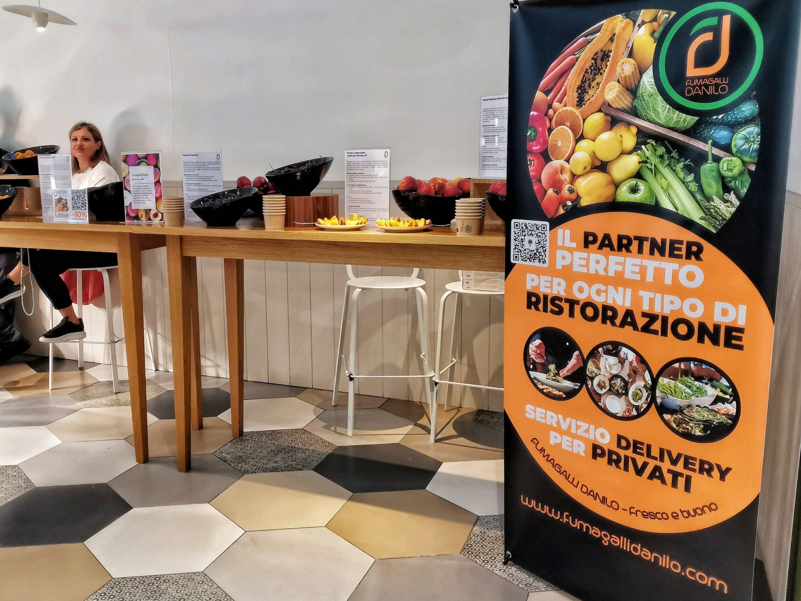 Fumagalli Danilo Ingrosso Ortofrutta Una donna è seduta a un tavolo alto su cui sono disposte delle ciotole di frutta; uno striscione colorato pubblicizza un servizio di vendita all’ingrosso di frutta e verdura per ristoranti e clienti privati. Ristorazione Professionale - GDO - retail - privati