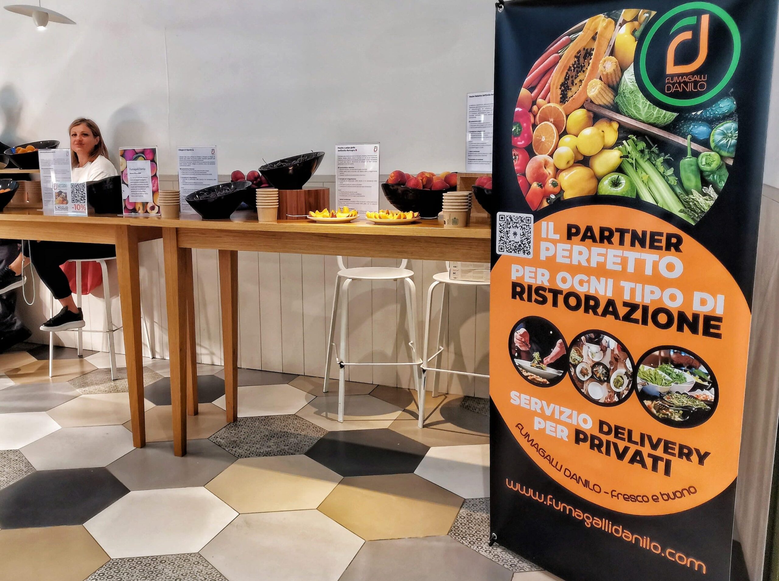 Fumagalli Danilo Ingrosso Ortofrutta Una donna è seduta a un tavolo alto su cui sono disposte delle ciotole di frutta; uno striscione colorato pubblicizza un servizio di vendita all’ingrosso di frutta e verdura per ristoranti e clienti privati. Ristorazione Professionale - GDO - retail - privati