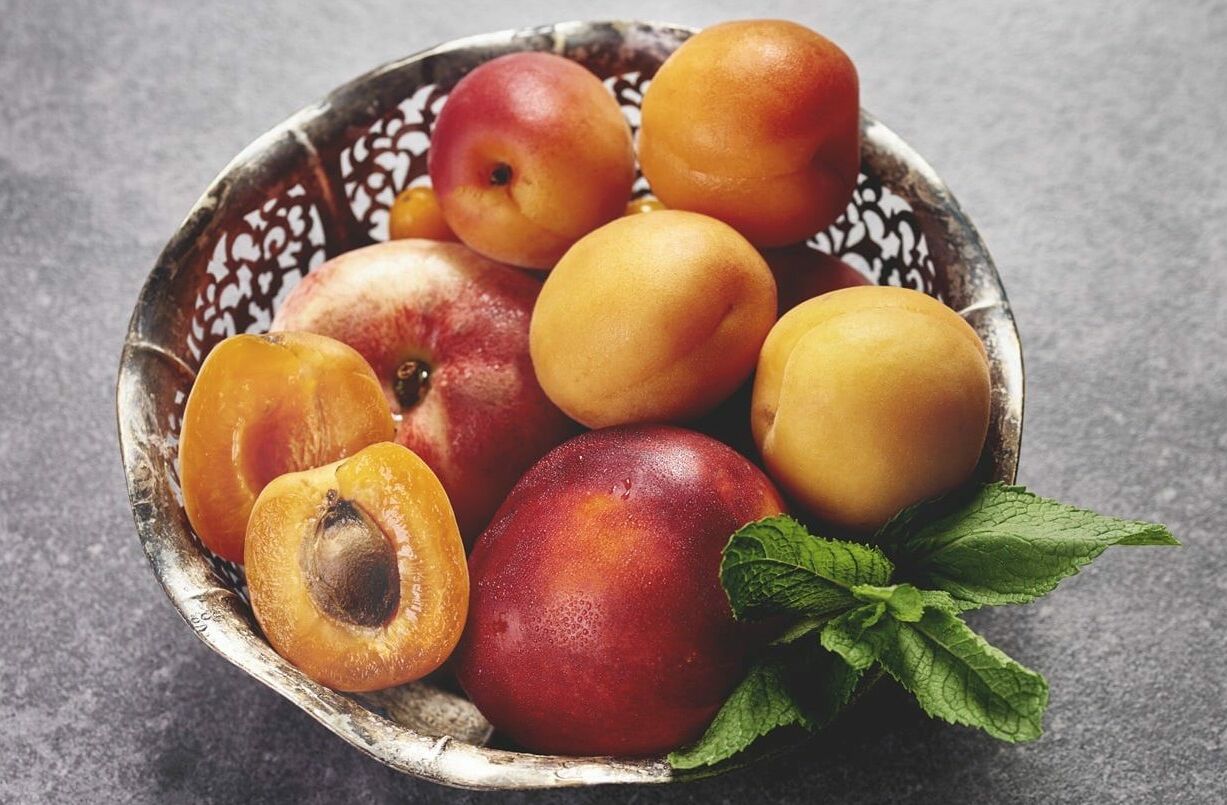 Fumagalli Danilo Ingrosso Ortofrutta Una ciotola di metallo piena di albicocche intere e tagliate a metà, una nettarina e un rametto di menta fresca, adagiata su una superficie grigia, mette in risalto la freschezza e la qualità che ci si aspetta da Fumagalli Danilo Besana in Brianza, il vostro grossista di ortofrutta di fiducia per la ristorazione. Ristorazione Professionale - GDO - retail - privati