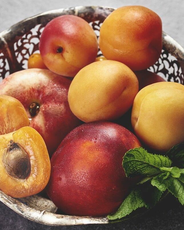 Fumagalli Danilo Ingrosso Ortofrutta Una ciotola di metallo piena di albicocche intere e tagliate a metà, una pesca e un rametto di menta fresca è appoggiata su una superficie grigia, a mettere in risalto la freschezza e la qualità che ci si aspetta da “ingrosso frutta e verdura” di Fumagalli Danilo Besana in Brianza. Ristorazione Professionale - GDO - retail - privati