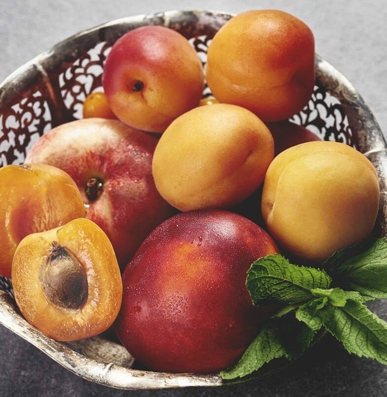 Fumagalli Danilo Ingrosso Ortofrutta Una ciotola di metallo piena di albicocche intere e tagliate a metà, una nettarina e un rametto di menta fresca, adagiata su una superficie grigia, mette in risalto la freschezza e la qualità che ci si aspetta da Fumagalli Danilo Besana in Brianza, il vostro grossista di ortofrutta di fiducia per la ristorazione. Ristorazione Professionale - GDO - retail - privati