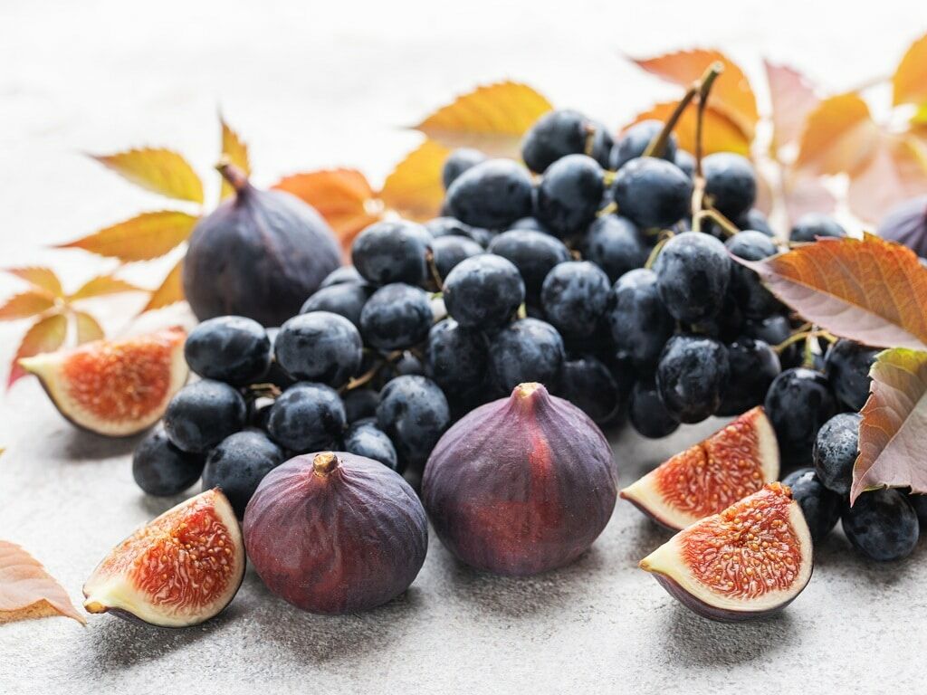 Fumagalli Danilo Ingrosso Ortofrutta Un grappolo d&rsquo;uva nera, fichi interi e fette di fico sono disposti su una superficie chiara, con foglie autunnali sparse tutt&rsquo;intorno, creando una composizione artistica ideale per la vendita all&rsquo;ingrosso di frutta e verdura o per la ristorazione ortofrutticola. Ristorazione Professionale - GDO - retail - privati