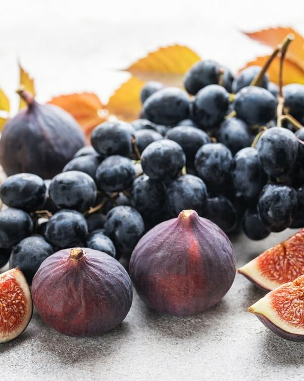 Fumagalli Danilo Ingrosso Ortofrutta Un grappolo d’uva nera, fichi interi e fette di fico sono disposti su una superficie chiara, con foglie autunnali sparse tutt’intorno, creando una composizione artistica ideale per la vendita all’ingrosso di frutta e verdura o per la ristorazione ortofrutticola. Ristorazione Professionale - GDO - retail - privati