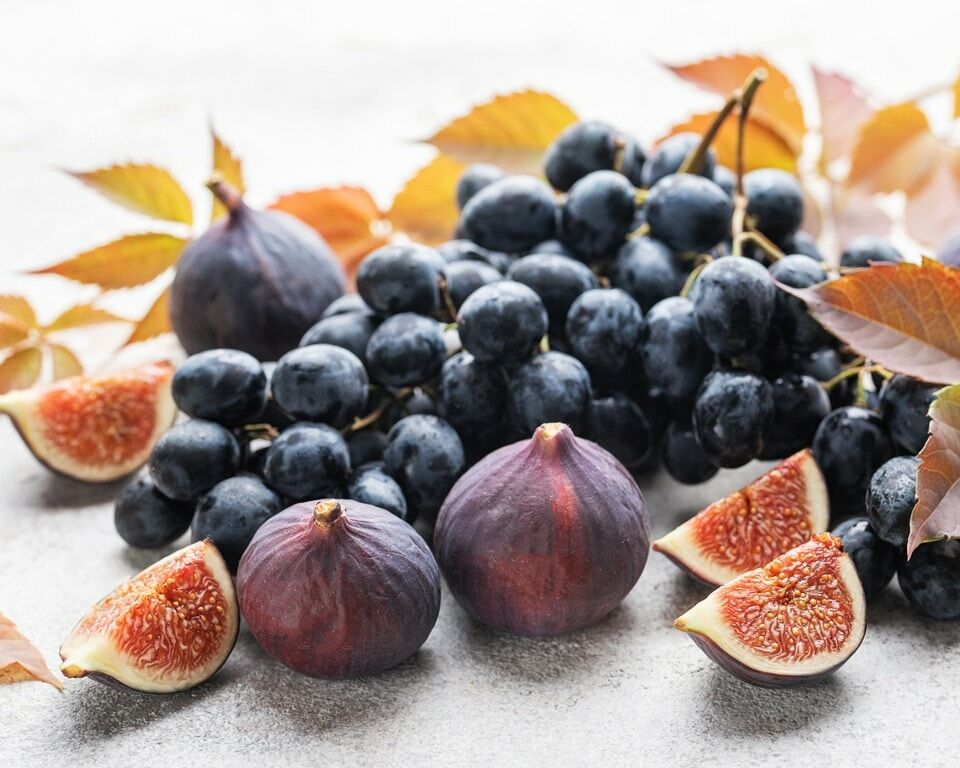 Fumagalli Danilo Ingrosso Ortofrutta Un grappolo d’uva nera, fichi interi e fette di fico sono disposti su una superficie chiara, con foglie autunnali sparse tutt’intorno, creando una composizione artistica ideale per la vendita all’ingrosso di frutta e verdura o per la ristorazione ortofrutticola. Ristorazione Professionale - GDO - retail - privati