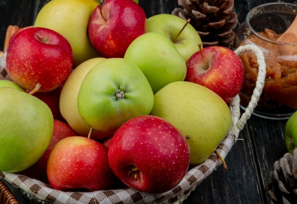 Fumagalli Danilo Ingrosso Ortofrutta Un cesto di vimini pieno di mele rosse e verdi, pigne e un barattolo di burro di mele con un cucchiaio di legno appoggiato su un piano in legno scuro: perfetto per mettere in risalto la freschezza che ci si aspetta da Fumagalli Danilo Besana in Brianza, il vostro grossista di fiducia per ortofrutta di qualità destinata alla ristorazione. Ristorazione Professionale - GDO - retail - privati