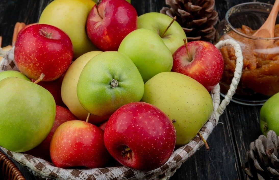 Fumagalli Danilo Ingrosso Ortofrutta Un cesto di vimini pieno di mele rosse e verdi, pigne e un barattolo di burro di mele con un cucchiaio di legno appoggiato su un piano in legno scuro: perfetto per mettere in risalto la freschezza che ci si aspetta da Fumagalli Danilo Besana in Brianza, il vostro grossista di fiducia per ortofrutta di qualità destinata alla ristorazione. Ristorazione Professionale - GDO - retail - privati
