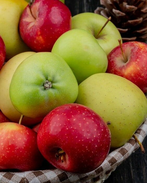 Fumagalli Danilo Ingrosso Ortofrutta Un cesto pieno di mele rosse e verdi, acquistato da un grossista di ortofrutta, è appoggiato su un piano di legno scuro, circondato da pigne e da un barattolo di confettura di mele. Ristorazione Professionale - GDO - retail - privati