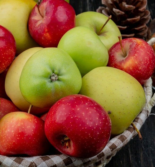 Fumagalli Danilo Ingrosso Ortofrutta Un cesto di vimini pieno di mele rosse e verdi, pigne e un barattolo di burro di mele con un cucchiaio di legno appoggiato su un piano in legno scuro: perfetto per mettere in risalto la freschezza che ci si aspetta da Fumagalli Danilo Besana in Brianza, il vostro grossista di fiducia per ortofrutta di qualità destinata alla ristorazione. Ristorazione Professionale - GDO - retail - privati