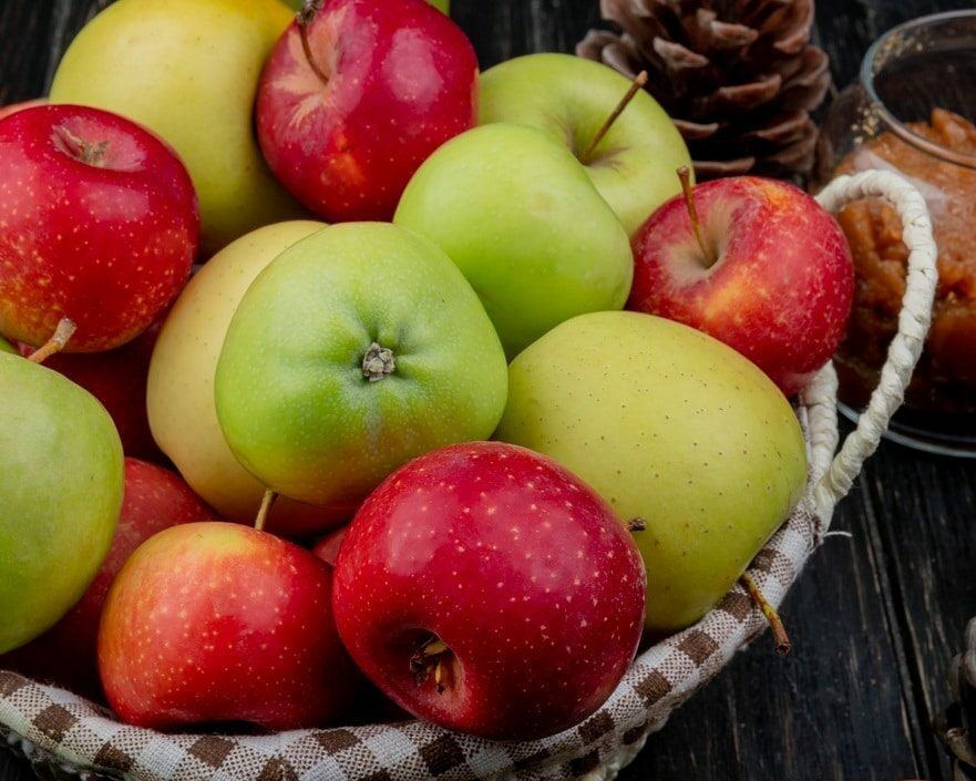 Fumagalli Danilo Ingrosso Ortofrutta Un cesto pieno di mele rosse e verdi, acquistato da un grossista di ortofrutta, è appoggiato su un piano di legno scuro, circondato da pigne e da un barattolo di confettura di mele. Ristorazione Professionale - GDO - retail - privati