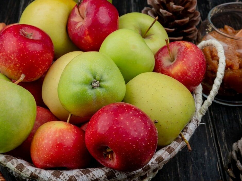 Fumagalli Danilo Ingrosso Ortofrutta Un cesto pieno di mele rosse e verdi, acquistato da un grossista di ortofrutta, è appoggiato su un piano di legno scuro, circondato da pigne e da un barattolo di confettura di mele. Ristorazione Professionale - GDO - retail - privati