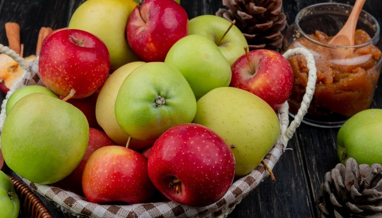 Fumagalli Danilo Ingrosso Ortofrutta Un cesto di vimini pieno di mele rosse e verdi, pigne e un barattolo di burro di mele con un cucchiaio di legno appoggiato su un piano in legno scuro: perfetto per mettere in risalto la freschezza che ci si aspetta da Fumagalli Danilo Besana in Brianza, il vostro grossista di fiducia per ortofrutta di qualità destinata alla ristorazione. Ristorazione Professionale - GDO - retail - privati