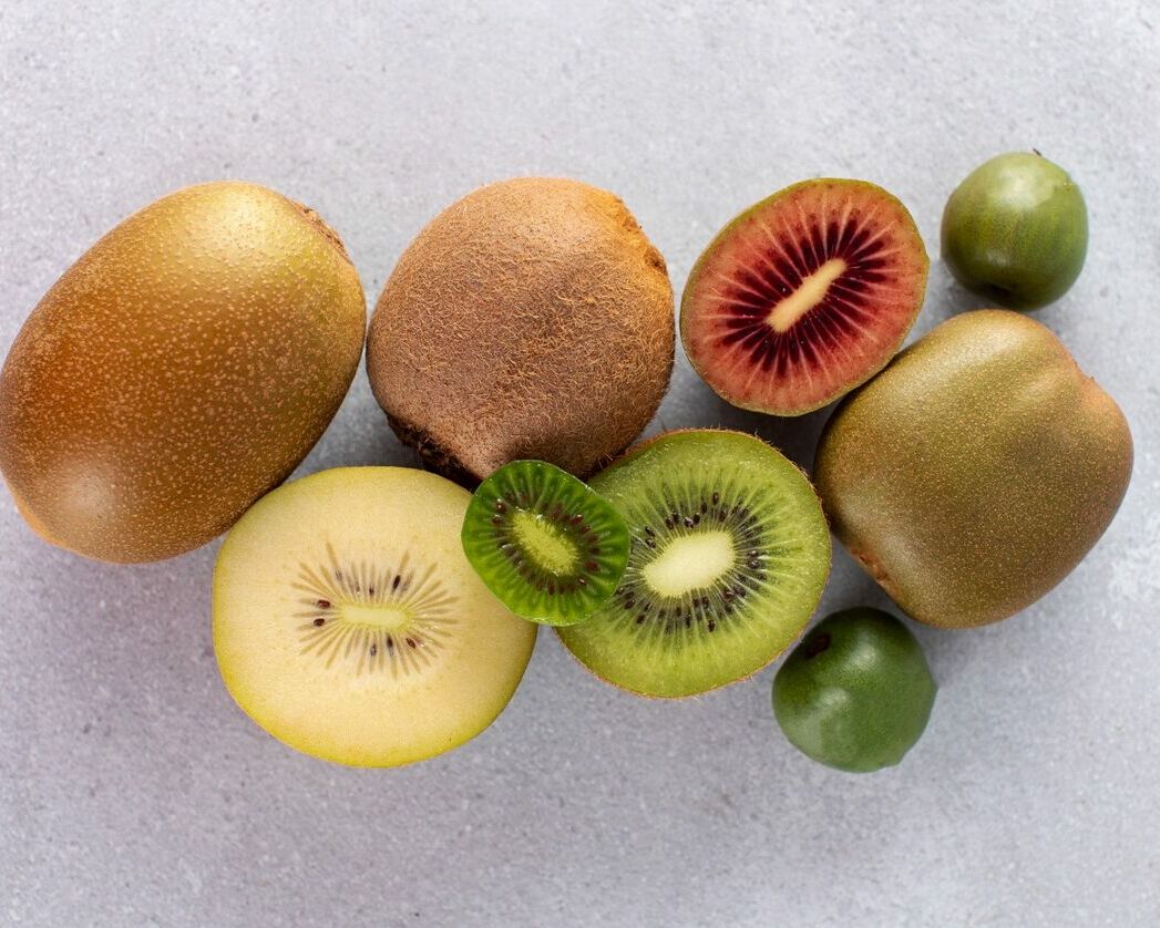 Fumagalli Danilo Ingrosso Ortofrutta Cinque kiwi, interi e a fette, di diverse varietà, con la polpa verde, gialla e rossa, sono disposti con gusto su una superficie grigio chiaro, mettendo in risalto la selezione di alta qualità offerta da Fumagalli Danilo Besana in Brianza, un affidabile grossista ortofrutticolo. Ristorazione Professionale - GDO - retail - privati