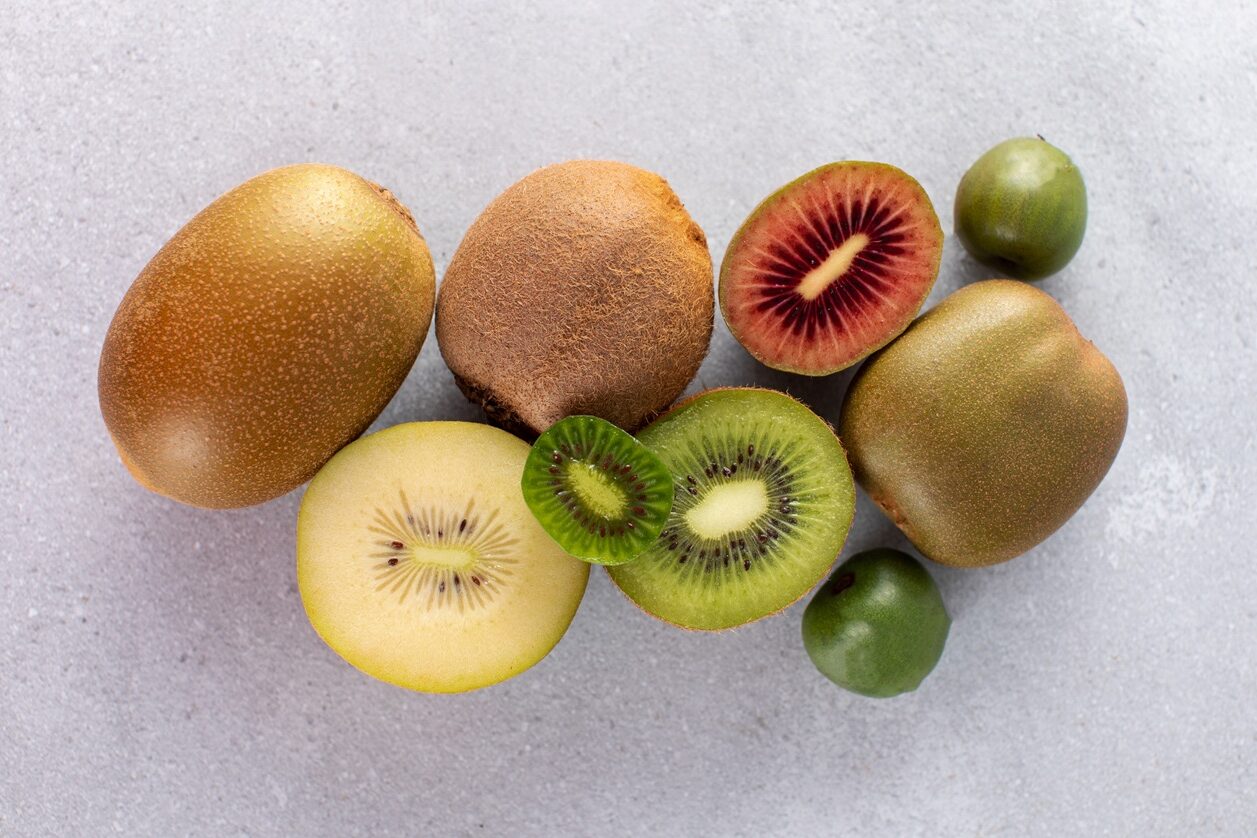 Fumagalli Danilo Ingrosso Ortofrutta Diversi kiwi interi e tagliati a metà, di diverse varietà, con la polpa verde, gialla e rossa, sono disposti su una superficie grigio chiaro da Fumagalli Danilo Besana in Brianza, uno dei principali grossisti ortofrutticoli. Ristorazione Professionale - GDO - retail - privati