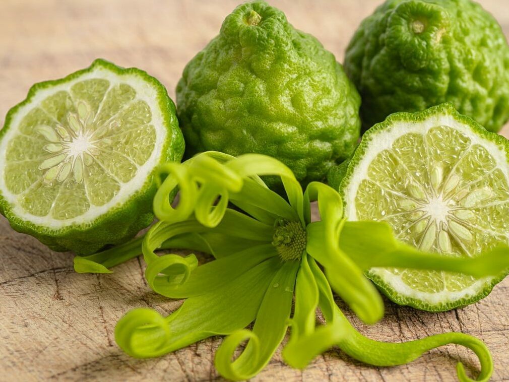 Fumagalli Danilo Ingrosso Ortofrutta Tre frutti di bergamotto, due dei quali tagliati a metà in modo da mostrarne l’interno, sono disposti su una superficie di legno con un fiore verde di ylang-ylang in primo piano: l’immagine perfetta per chi è alla ricerca di ingredienti di qualità presso un grossista ortofrutta o presso aziende specializzate nel commercio all’ingrosso di frutta e verdura. Ristorazione Professionale - GDO - retail - privati