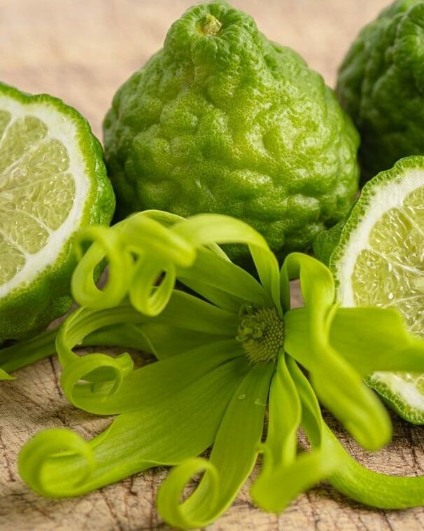 Fumagalli Danilo Ingrosso Ortofrutta Tre frutti di bergamotto, due dei quali tagliati a metà in modo da mostrarne l’interno, sono disposti su una superficie di legno con un fiore verde di ylang-ylang in primo piano: l’immagine perfetta per chi è alla ricerca di ingredienti di qualità presso un grossista ortofrutta o presso aziende specializzate nel commercio all’ingrosso di frutta e verdura. Ristorazione Professionale - GDO - retail - privati