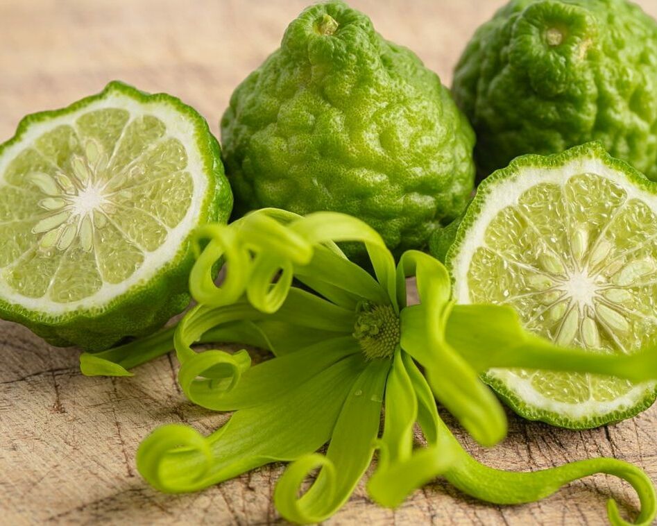 Fumagalli Danilo Ingrosso Ortofrutta Tre frutti di bergamotto, due dei quali tagliati a metà in modo da mostrarne l’interno, sono disposti su una superficie di legno con un fiore verde di ylang-ylang in primo piano: l’immagine perfetta per chi è alla ricerca di ingredienti di qualità presso un grossista ortofrutta o presso aziende specializzate nel commercio all’ingrosso di frutta e verdura. Ristorazione Professionale - GDO - retail - privati