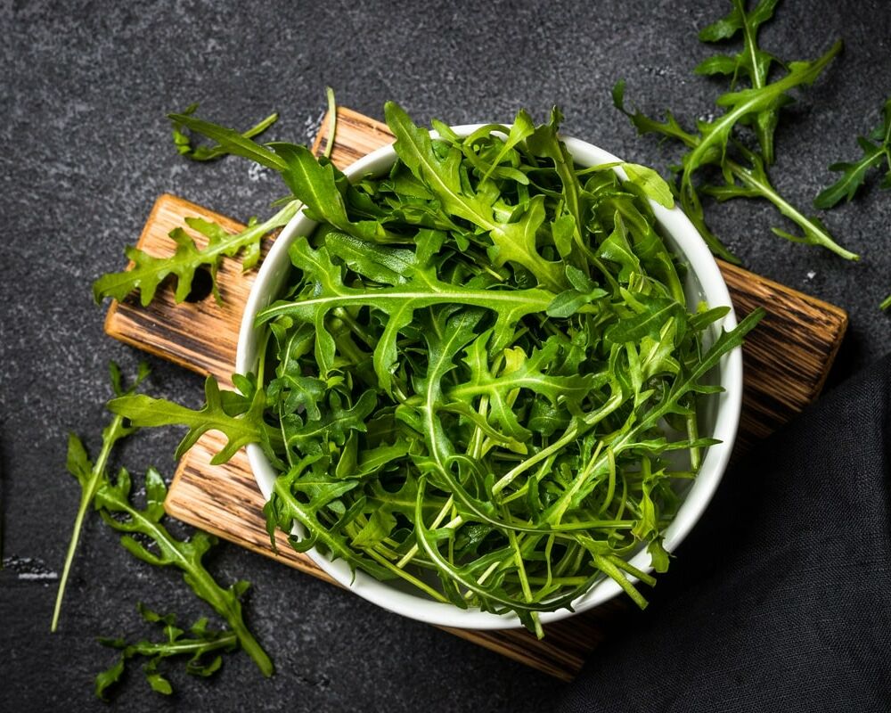 Fumagalli Danilo Ingrosso Ortofrutta Una ciotola bianca piena di foglie fresche di rucola provenienti da Fumagalli Danilo Besana in Brianza è appoggiata su un tagliere di legno, con altra rucola proveniente da questo affidabile grossista ortofrutticolo sparsa su una superficie scura. Ristorazione Professionale - GDO - retail - privati
