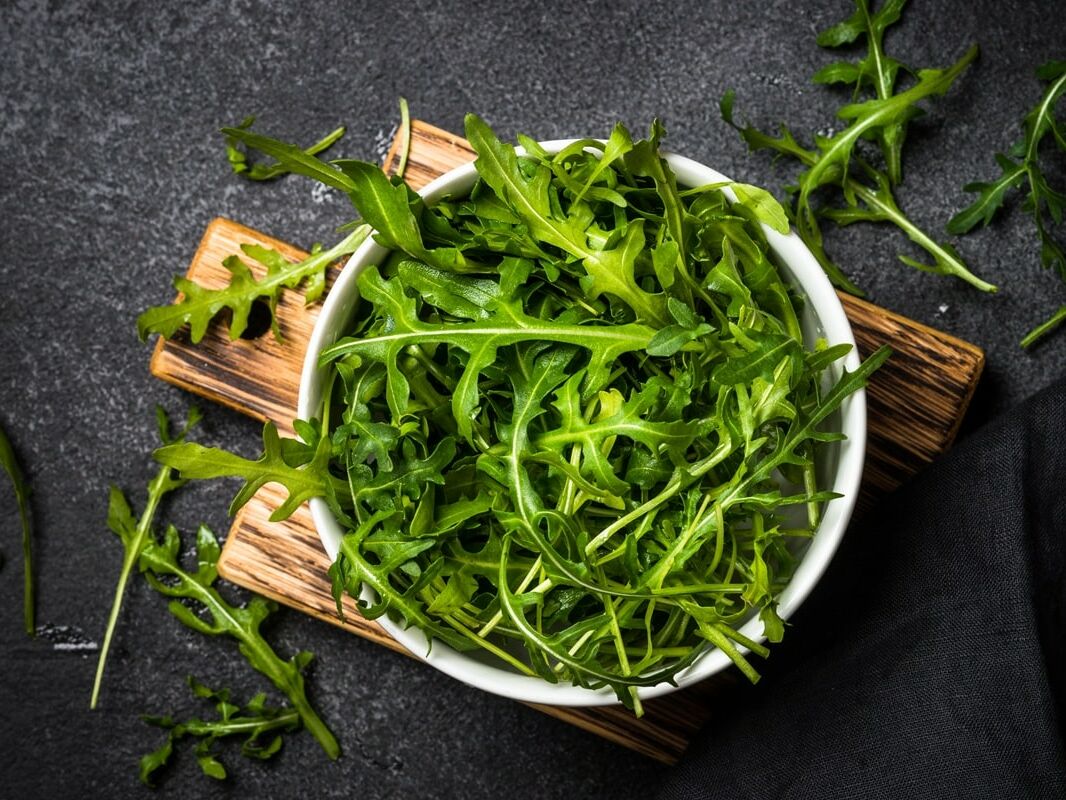 Fumagalli Danilo Ingrosso Ortofrutta Una ciotola bianca piena di foglie fresche di rucola di Fumagalli Danilo Besana in Brianza è appoggiata su un tagliere di legno, con alcune foglie sparse su una superficie scura. Ristorazione Professionale - GDO - retail - privati