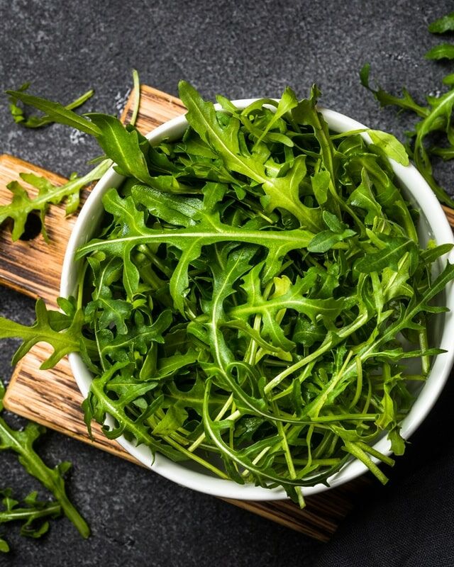 Fumagalli Danilo Ingrosso Ortofrutta Una ciotola bianca piena di foglie fresche di rucola provenienti da Fumagalli Danilo Besana in Brianza è appoggiata su un tagliere di legno, con altra rucola proveniente da questo affidabile grossista ortofrutticolo sparsa su una superficie scura. Ristorazione Professionale - GDO - retail - privati