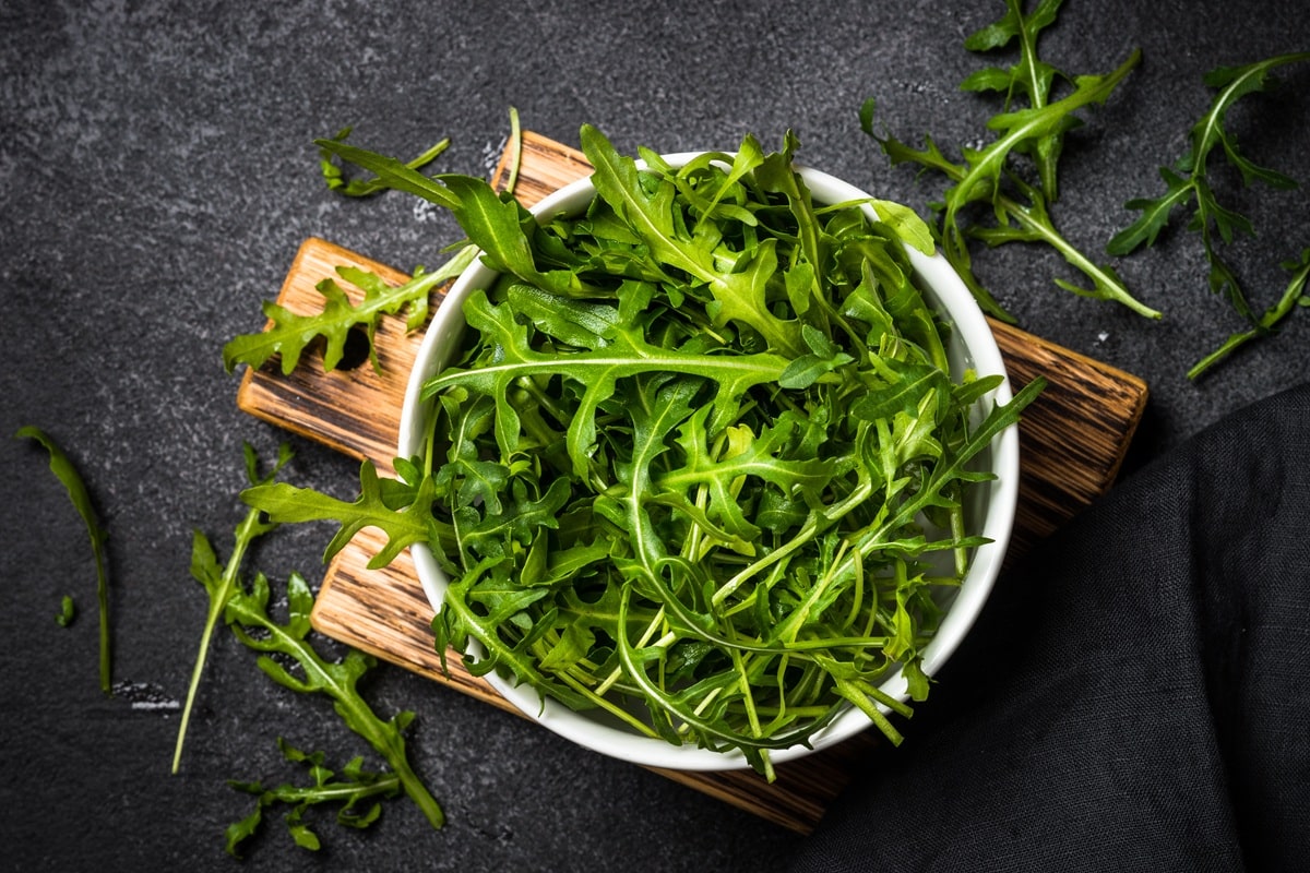 Fumagalli Danilo Ingrosso Ortofrutta Una ciotola bianca piena di foglie fresche di rucola di Fumagalli Danilo Besana in Brianza è appoggiata su un tagliere di legno, con alcune foglie sparse su una superficie scura. Ristorazione Professionale - GDO - retail - privati