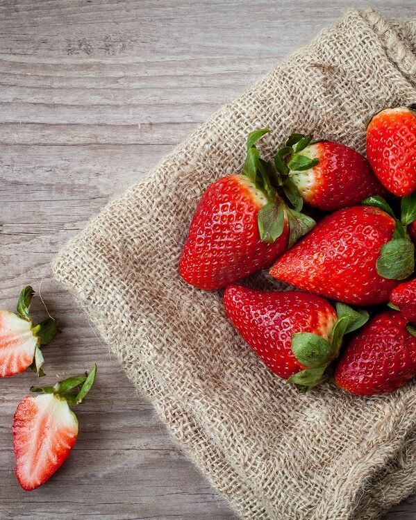 Fumagalli Danilo Ingrosso Ortofrutta Un gruppo di fragole intere è disposto su un telo di juta, con due fragole affettate poste accanto su una superficie di legno, a testimonianza della qualità che contraddistingue Fumagalli Danilo Besana in Brianza e della sua competenza nel settore della vendita all’ingrosso di frutta e verdura. Ristorazione Professionale - GDO - retail - privati