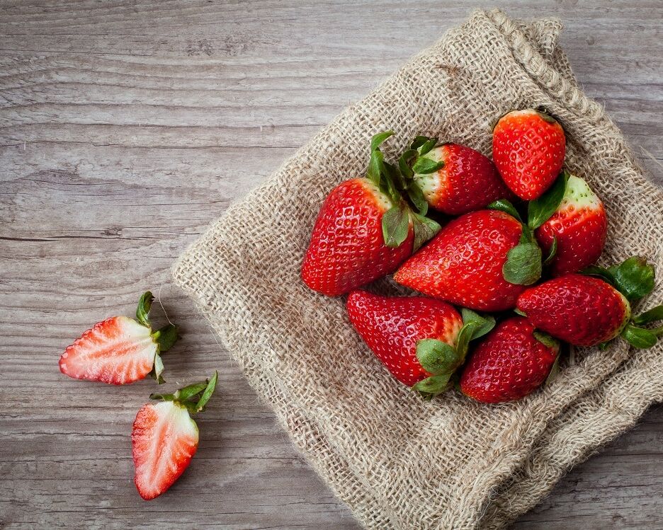 Fumagalli Danilo Ingrosso Ortofrutta Un gruppo di fragole intere è disposto su un telo di juta, con due fragole affettate poste accanto su una superficie di legno, a testimonianza della qualità che contraddistingue Fumagalli Danilo Besana in Brianza e della sua competenza nel settore della vendita all’ingrosso di frutta e verdura. Ristorazione Professionale - GDO - retail - privati