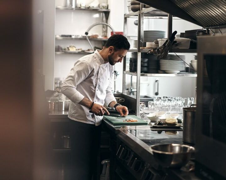 Fumagalli Danilo Ingrosso Ortofrutta Uno chef in giacca bianca taglia gli ingredienti acquistati dal grossista ortofrutta su un tagliere verde in una cucina professionale, circondato da utensili, stoviglie e attrezzature da cucina. Ristorazione Professionale - GDO - retail - privati
