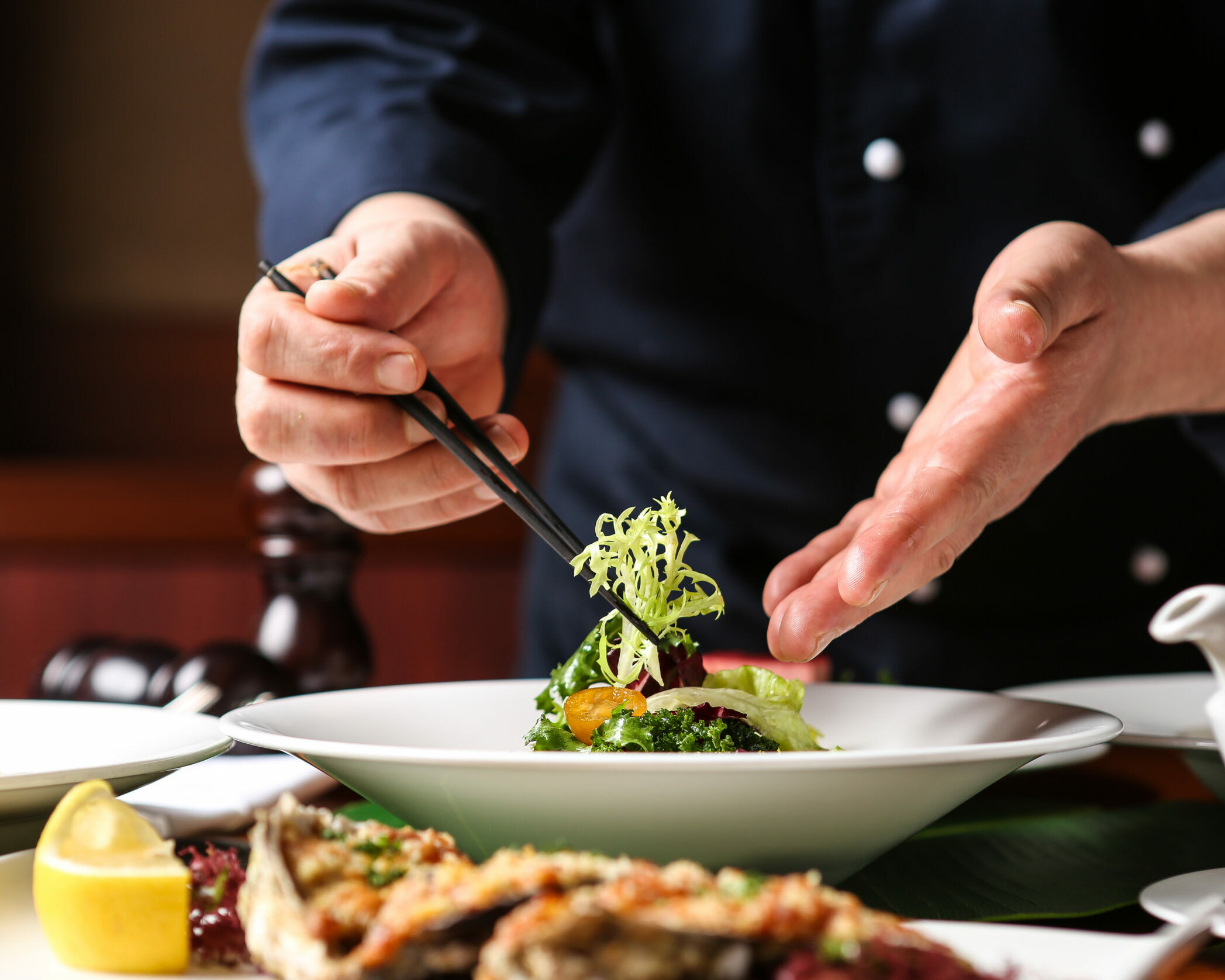 Fumagalli Danilo Ingrosso Ortofrutta Uno chef guarnisce un'insalata in una ciotola bianca con delle pinzette, mentre in primo piano si vedono altri piatti già impiattati e una fetta di limone, preparati con ingredienti di qualità