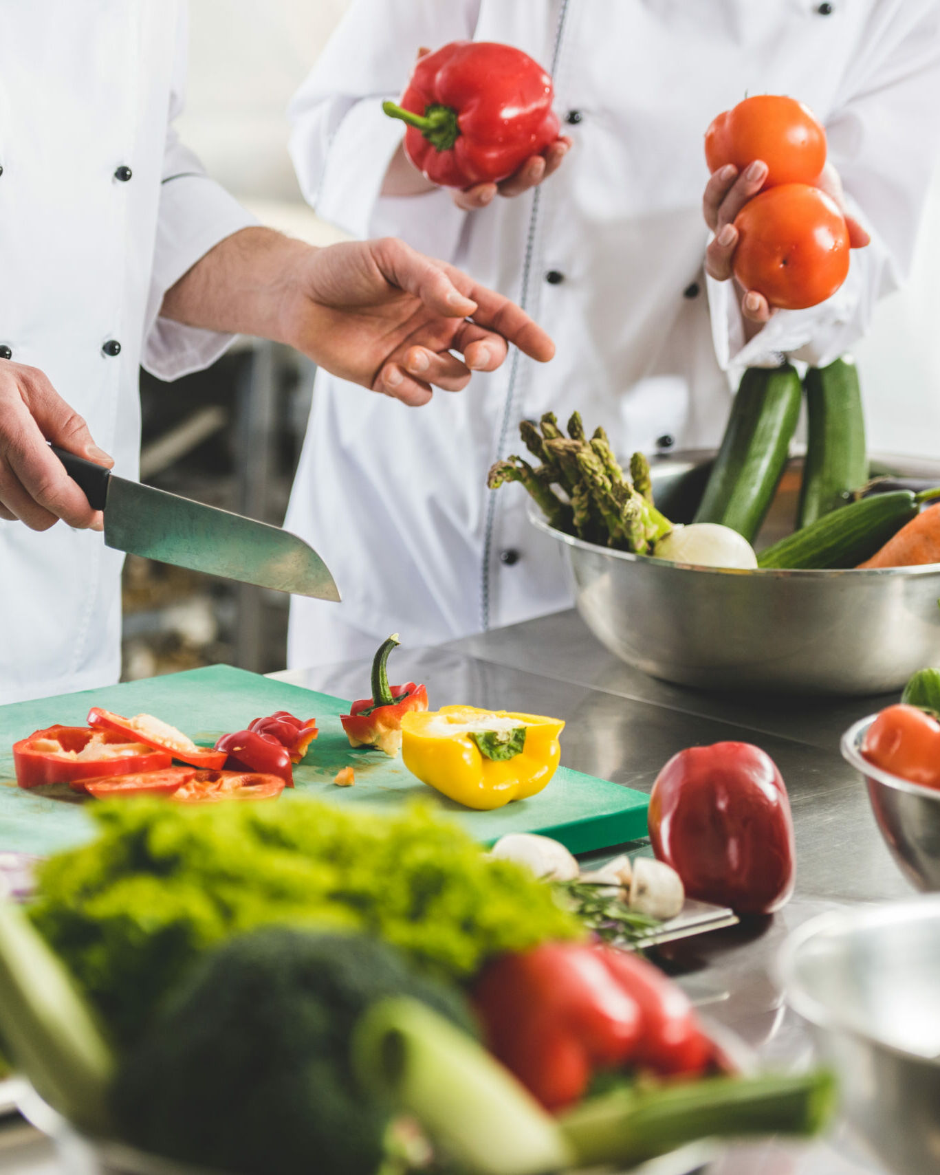 Fumagalli Danilo Ingrosso Ortofrutta Due cuochi in divisa bianca preparano e tagliano verdure fresche di vario tipo, tra cui peperoni, provenienti da un grossista ortofrutta, su un piano di lavoro in acciaio inossidabile. Ristorazione Professionale - GDO - retail - privati
