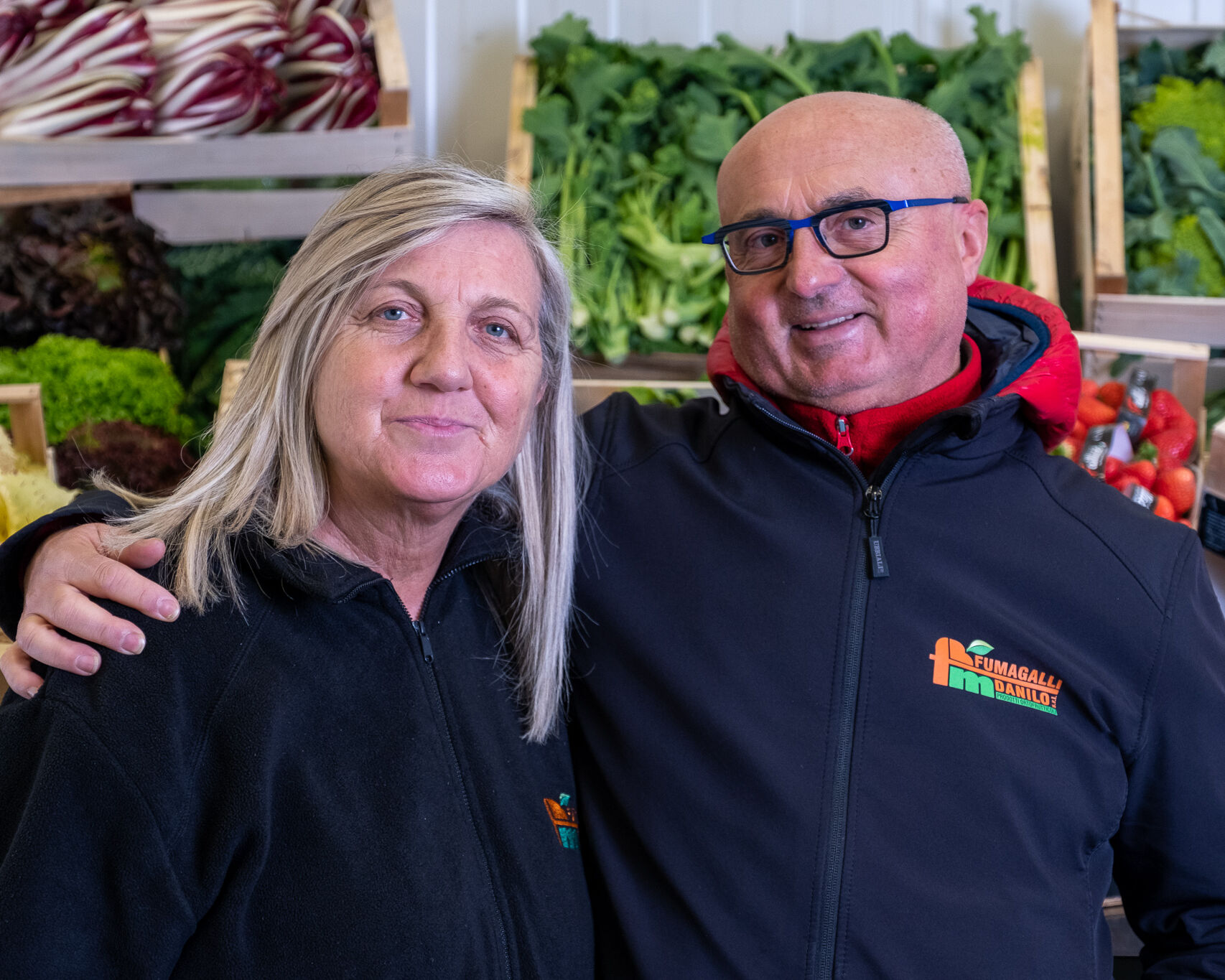 Fumagalli Danilo Ingrosso Ortofrutta Due persone con giacche nere stanno in piedi una accanto all&rsquo;altra e sorridono davanti a cassette di verdura piene di verdure a foglia verde, radicchio e fragole all&rsquo;interno di un mercato specializzato nella vendita all&rsquo;ingrosso di frutta e verdura e nella fornitura di ortofrutta per la ristorazione. Ristorazione Professionale - GDO - retail - privati