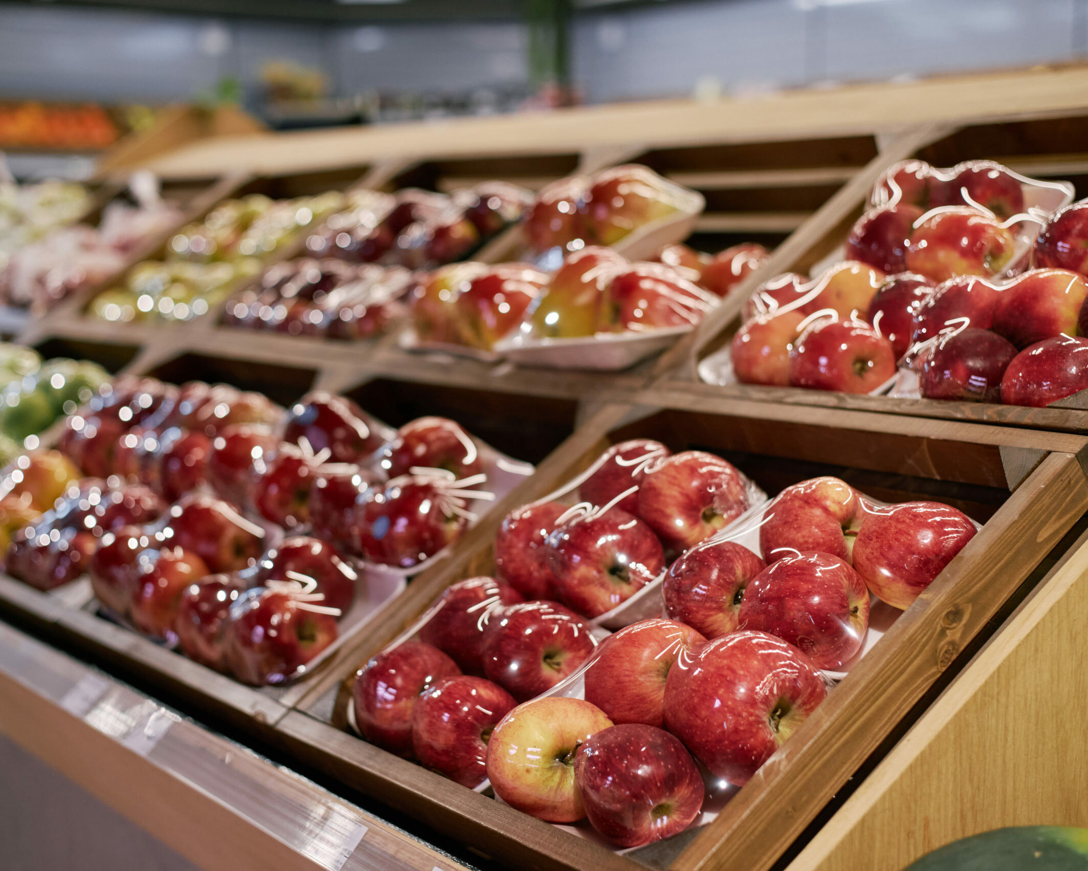 Fumagalli Danilo Ingrosso Ortofrutta Le mele avvolte nella pellicola trasparente e disposte su vassoi di legno sono esposte su ripiani inclinati nel reparto ortofrutta di un supermercato, a testimonianza della cura nella selezione tipica dei fornitori all’ingrosso di frutta e verdura come Fumagalli Danilo Besana in Brianza. Ristorazione Professionale - GDO - retail - privati